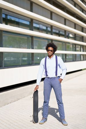 Man in sunglasses standing tall with skateboard outside building