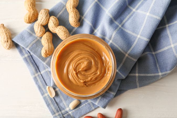 Glass jar with peanut butter, peanut and kitchen towel on wooden background, space for text and top view