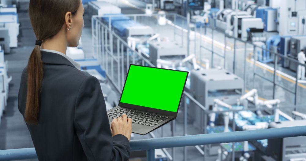 Female Technician Using Laptop Computer with Green Screen Chroma Key Mock Up Display and Looking Around a Factory Facility with Robotic Arms Producing Modern Electronic Components for Tech Industries