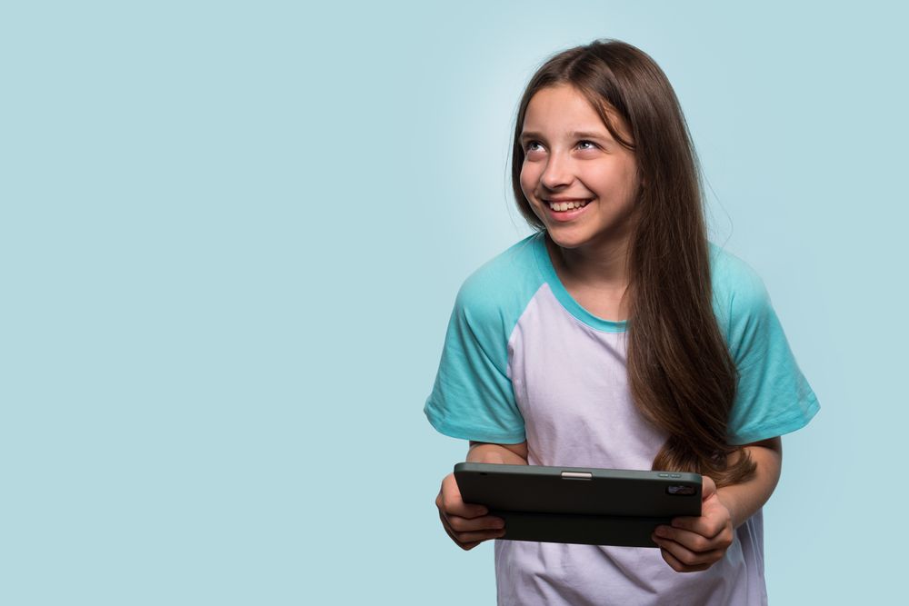 Cheerful girl looking upward, holding tablet in relaxed stance.