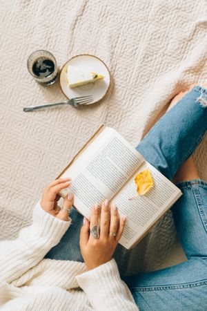 Top view of woman in denim pants reading a book in bed beside a cake