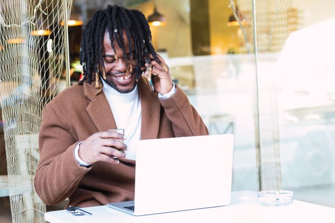 Cheerful Black man with laptop speaking on phone on restaurant terrace
