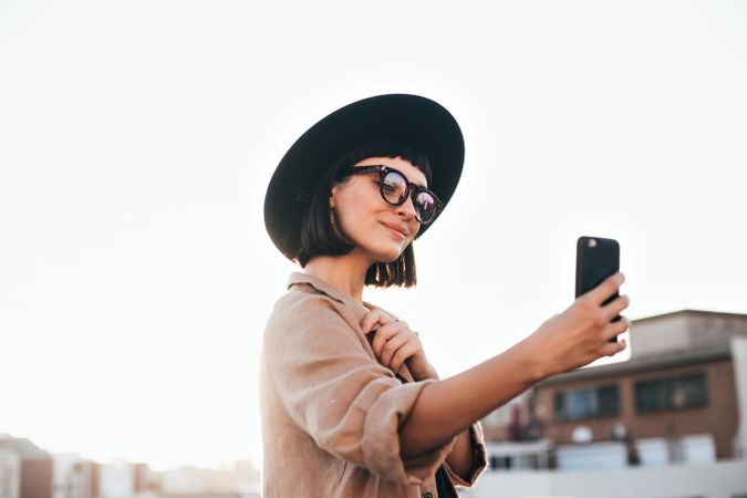Fashionable woman in thick glasses and hat taking selfie outside