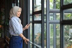Asian Muslim Businesswoman in Hijab  Holding Smartphone while Standing by Window