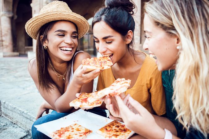 Three cheerful multi-ethnic young women eating Italian pizza at city street