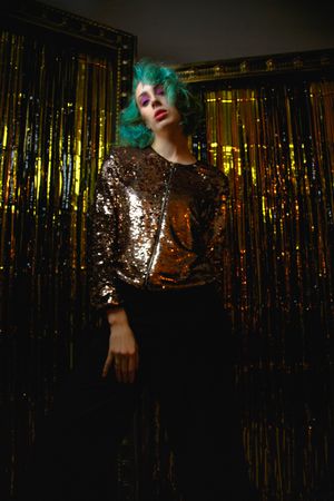 Woman with green hair and golden jacket standing beside curtain