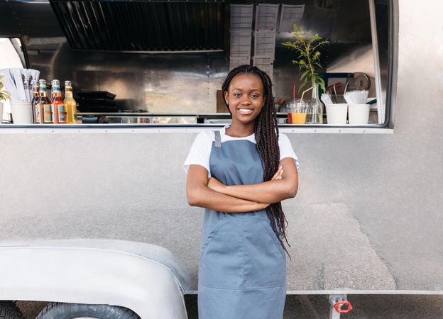 Proud Black woman in apron outside her silver food truck with arms crossed