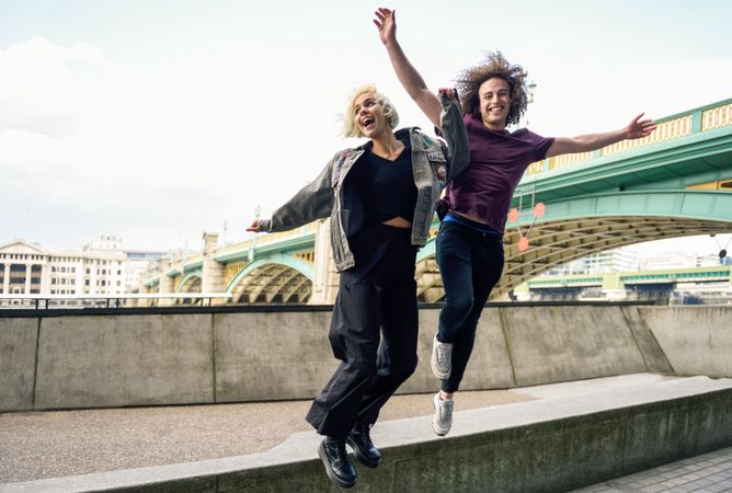 Male and female jumping and smiling on riverwalk with bridge in background