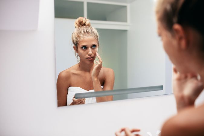 Woman applying cosmetic cream on face standing in the bathroom