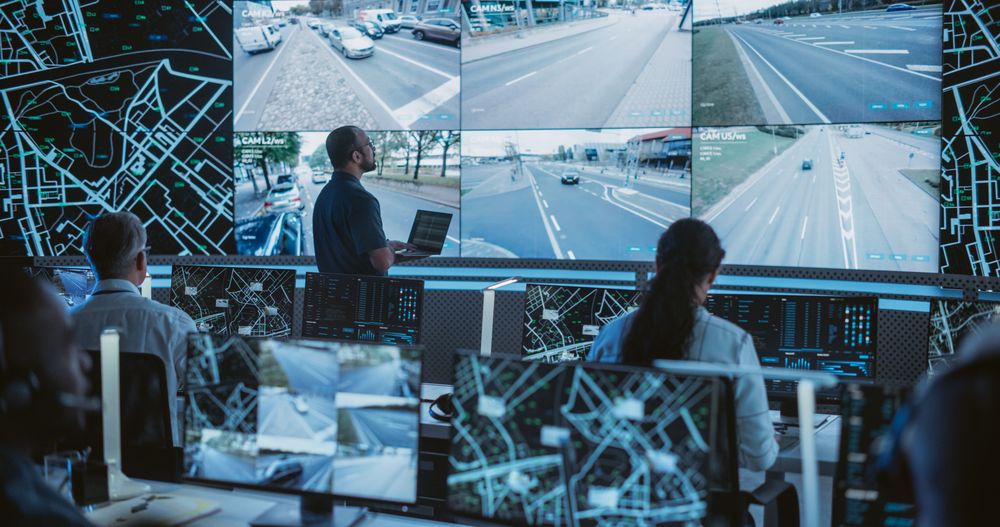 Male Software Engineers Working on Laptop Next to a Big Screen in a Modern Monitoring Office with Live CCTV Footage with Traffic Situation.