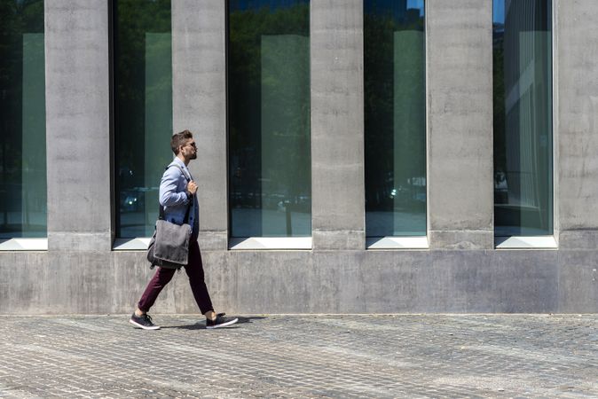 Side view of man walking in the sun with bag outside of building