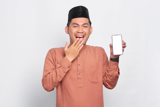 Muslim man in kufi hat with hand over mouth smiling with mobile phone
