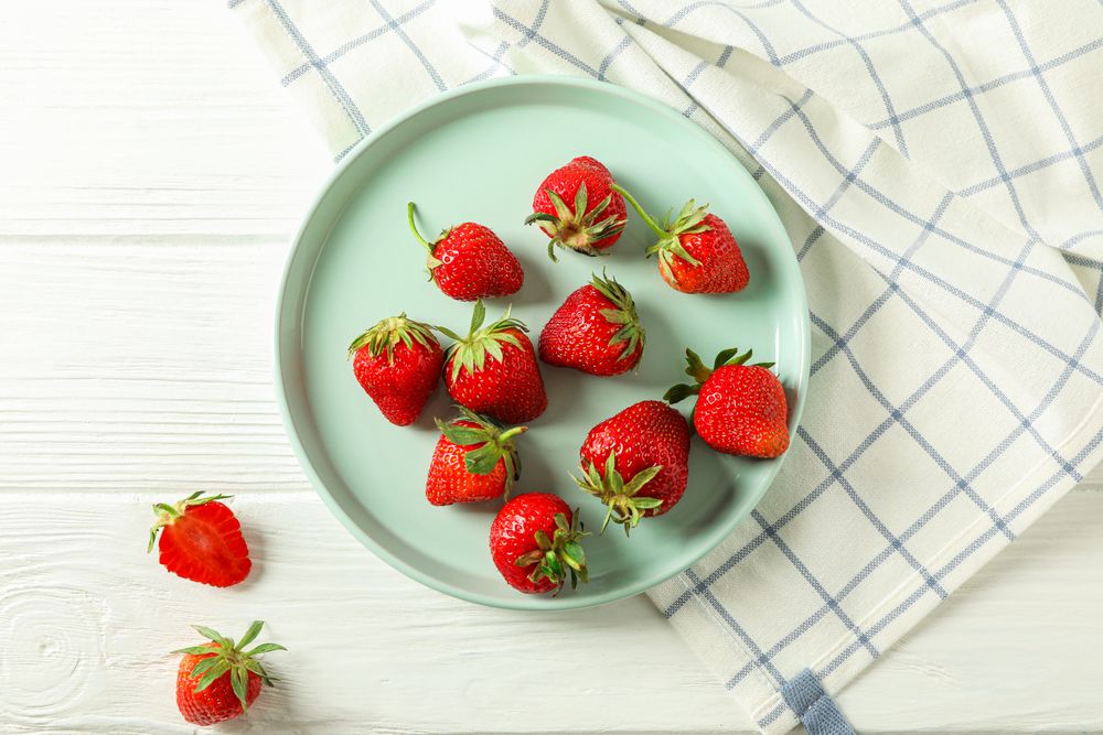 Plate with fresh strawberries and kitchen towel on bright wooden background, top view and space for text