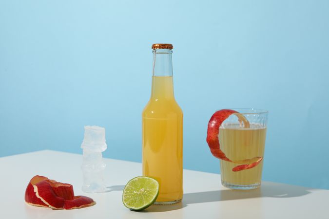 Glass and bottle with apple cider, piece of lime, ice cubes and apple peels on blue background