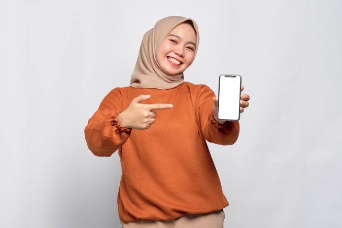 Smiling Muslim woman smiling holding smart phone and pointing finger at mockup screen