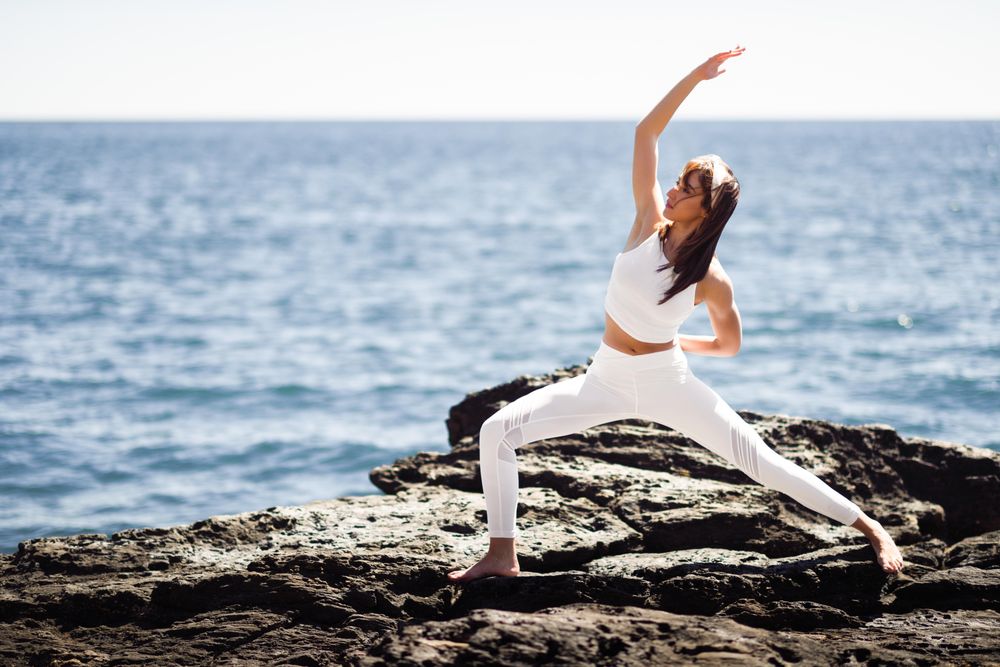 Woman wearing sport clothes and doing warrior A pose on rocks on the coast