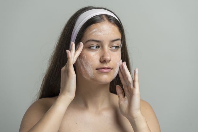 Beautiful woman looking at camera while washing her face with a cleansing scrub or mask