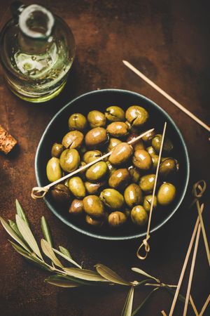 Bowl of olives on wooden table with garnish and bottle of olive oil, and skewers for serving