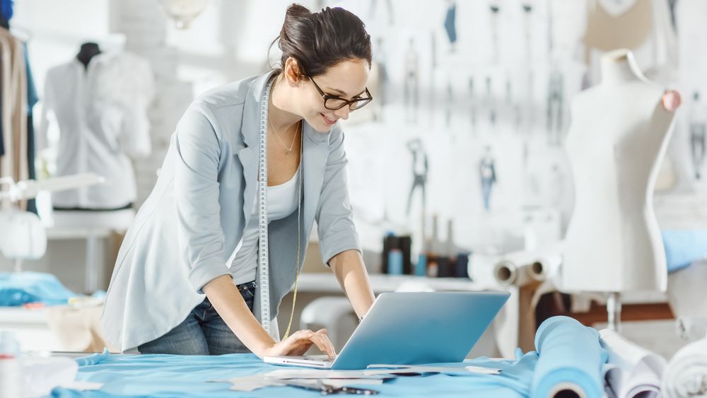 Female Designer Uses Laptop on Her Desk that is Covered with Various Fabrics and Sewing Items.