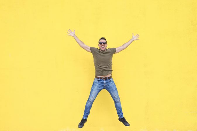 Happy male standing outside jumping in front of yellow wall with open arms