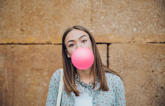 Female standing outside in front of stone wall blowing bubble with pink gum