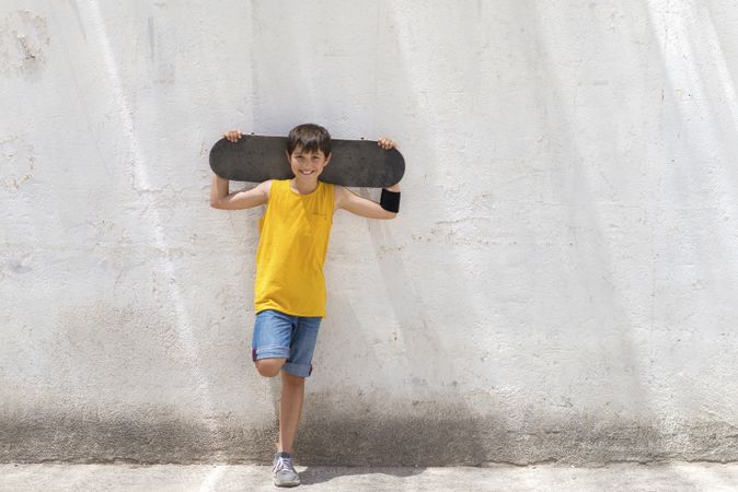 Young smiling boy leaning on wall holding a skateboard while looking camera on sunny day