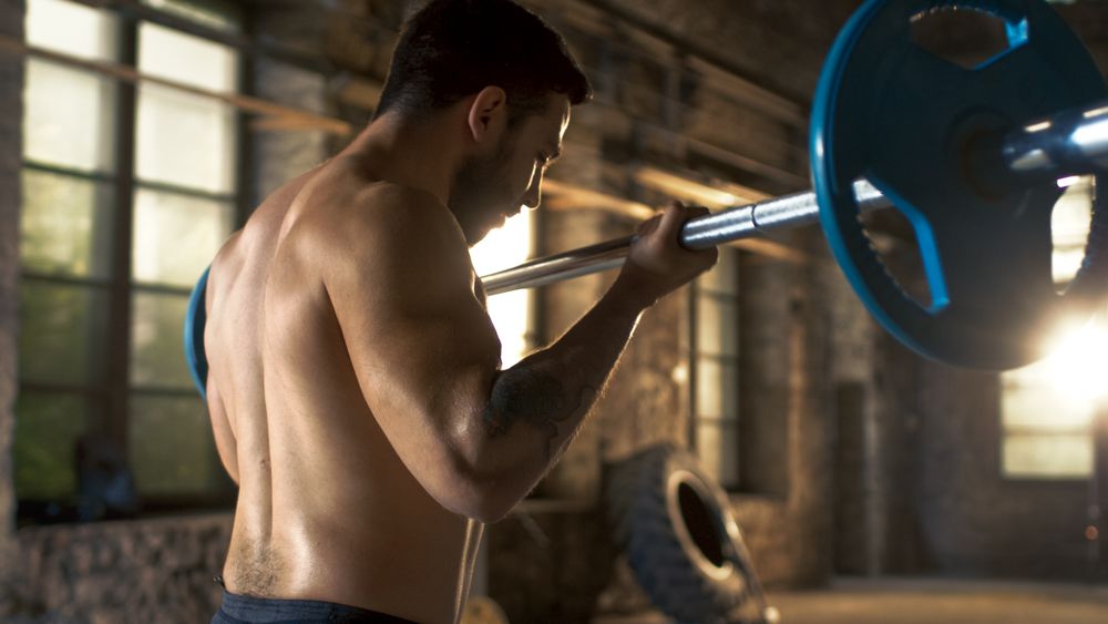 Muscular Shirtless Man Lifting Heavy Barbell with Effort, Doing Biceps Curls  Bodybuilding Exercise in the Industrial Gym Building.