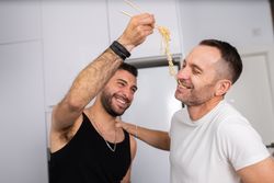 Laughing male couple playfully tasting noodles as they prepare meal