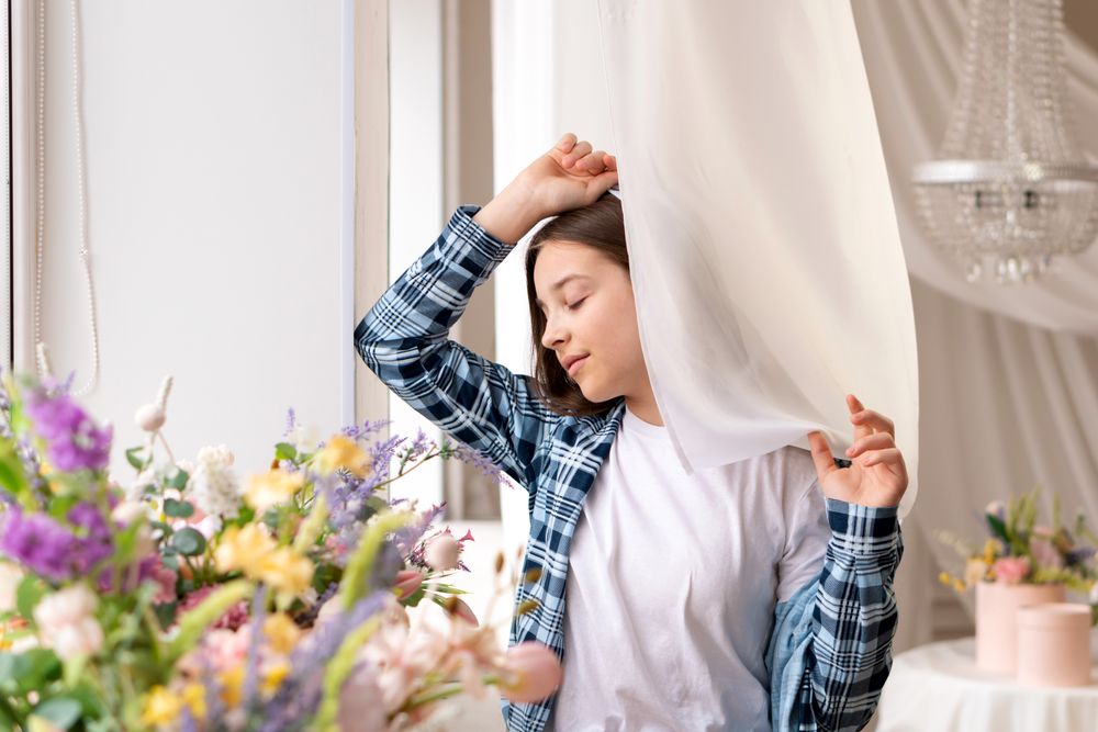 Girl holding curtain thoughtfully looking outside window near flowers.