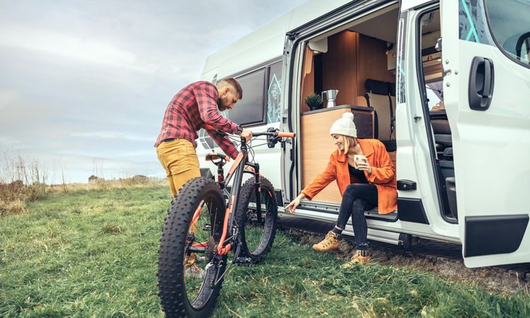Female in orange sipping coffee sitting in camper van with male friend tending to bicycle