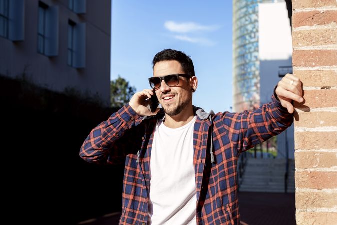 Bearded man standing by brick wall wearing sunglasses and chatting on smartphone outdoors