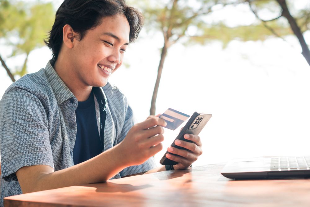 Happy Asian Young businessman hold blue creditcard and phone purchasing online shopping in outdoor cafe.