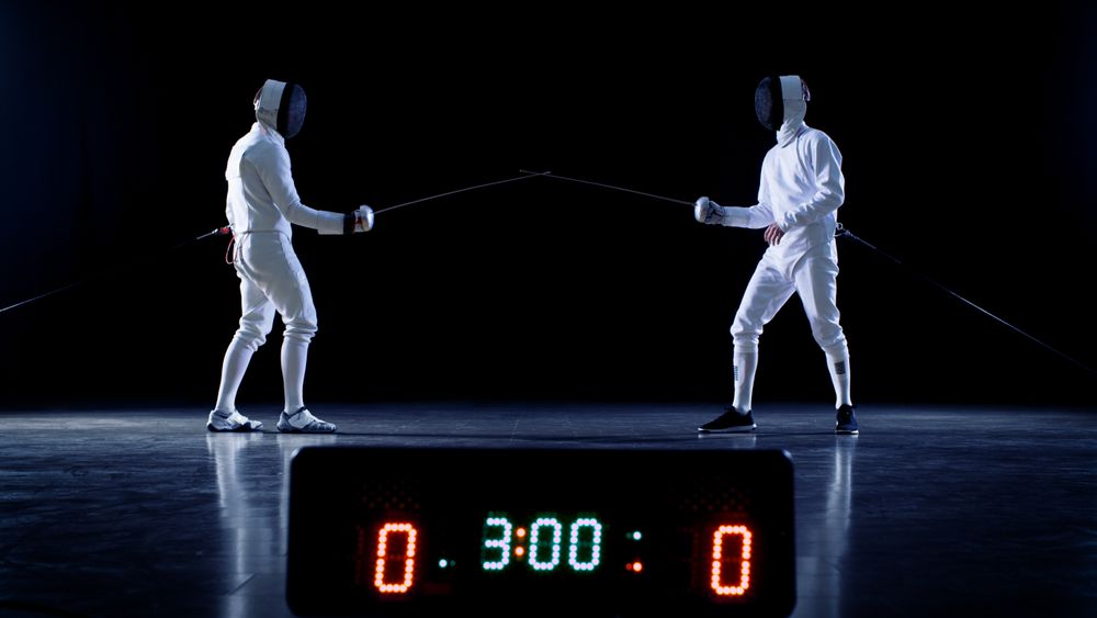 On the Championship Two Professional Fully Equipped Fencers Ready for Fight with Foils while Scoreboard Keeps the Scores of Hits.