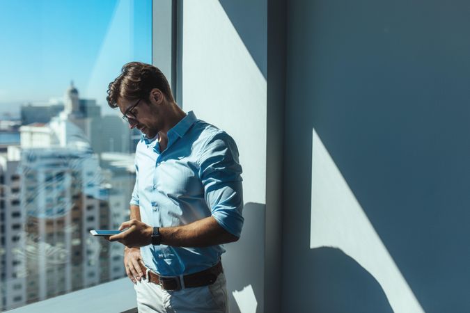 Professional male looking at cell phone next to glass window in office