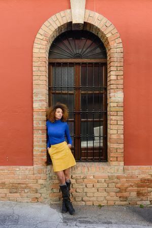 Nonchalant female with curly hair wearing bright blue shirt and yellow skirt in front of window