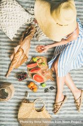 Woman lounging on picnic blanket with large hat thatched bag with glass of rose, baguette, fruit 4OEQL0