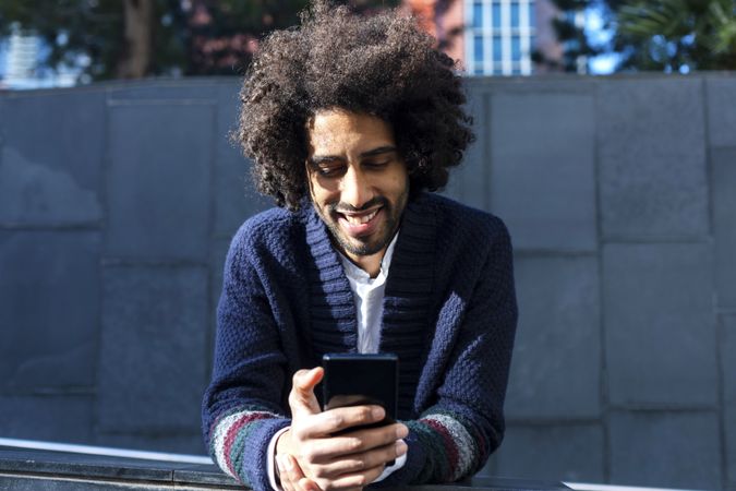 Black man in cardigan leaning forward outdoors on sunny day texting on smartphone