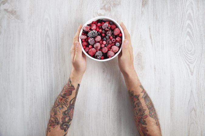 Arms of tattooed man holding ceramic bowl of fruit