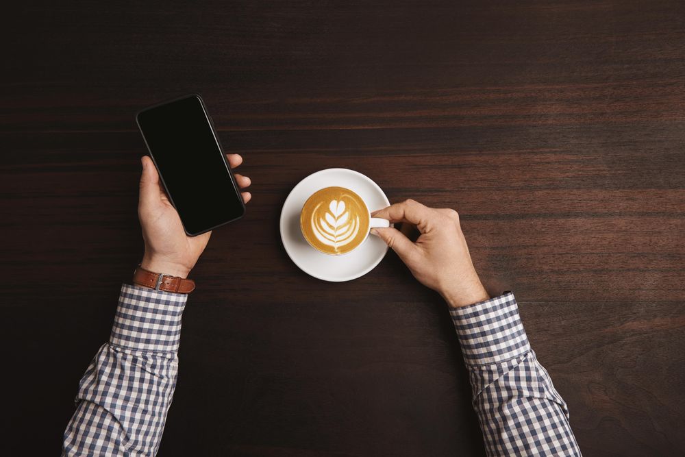 Arms in plaid shirt and leather wrist watch with cappuccino and cell phone