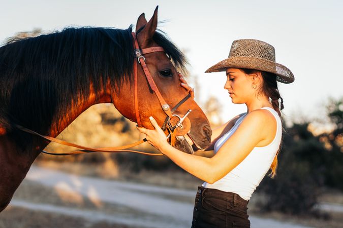 Young woman in hat walking with her horse in the countryside at sunset