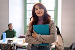 Cheerful young woman clutching a blue notebook in a classroom setting and walking