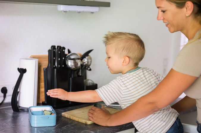 Happy crop mother and kid son with chopping board in kitchen in daylight