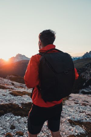 Back view of a man in red jacket with backpack hiking on shrubland at sunset