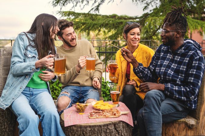 Four adults sit outdoors around a tree trunk table, sharing laughs and beers