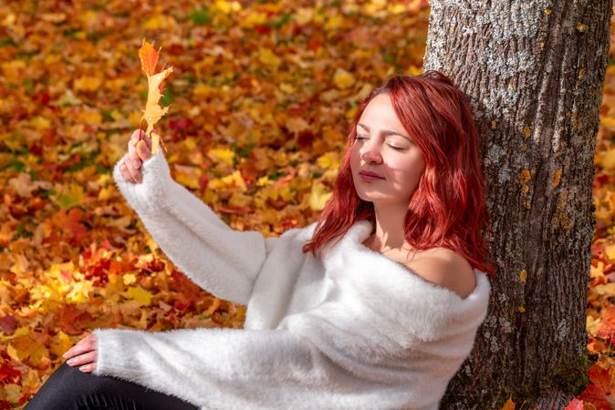 Woman leaning on tree holding up autumn leaf creating shadow on face