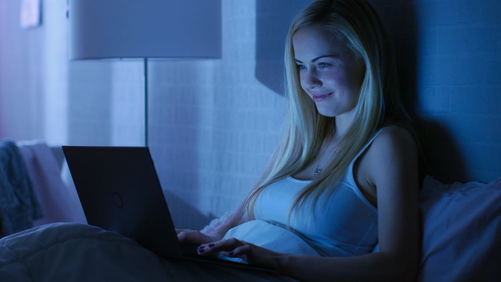 Young Woman Lies in Bed at Night and Works On Her Laptop.