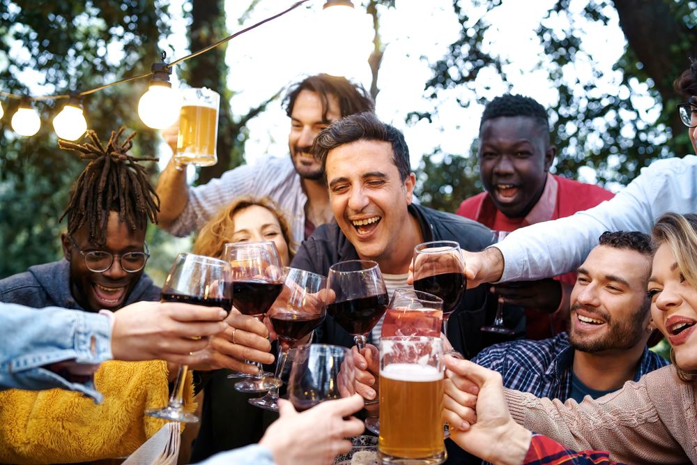 A diverse group of friends aged 20-40, toasting with beer, wine, and sangria glasses outdoors at a countryside house during dusk