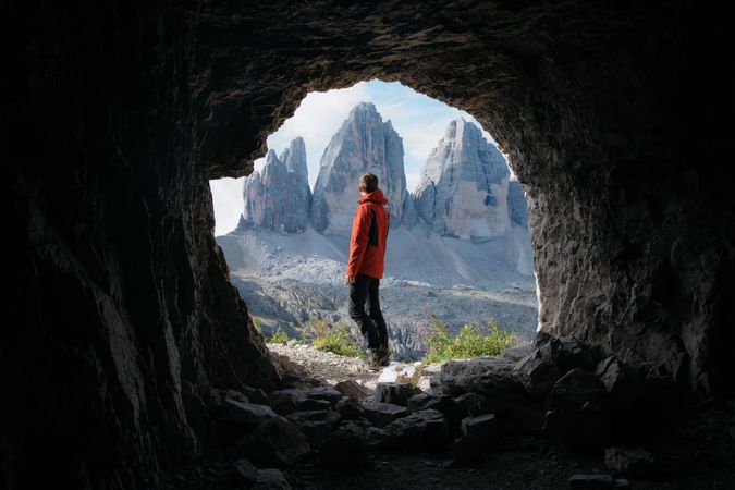 Man in red jacket standing beside cave near mountains