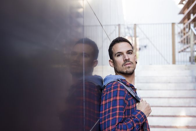 Man in plaid shirt leaning on shiny wall outdoors in front of stairs on a sunny day
