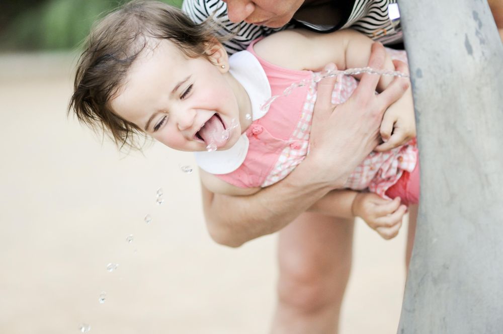 Little girl drinking water from fountain in her mother’s arms